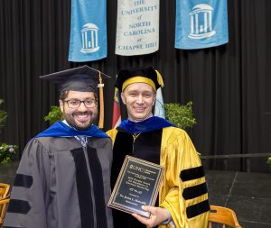 Two men are smiling while posing in front of the stage at the doctoral hooding ceremony. The man on the right wears gold colored academic robes and is holding a plaque. The man on the left is in black academic robes.