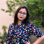 Sampreeti Bhattacharya wearing a polkadot shirt and glasses. She is standing with her hand on her hip.
