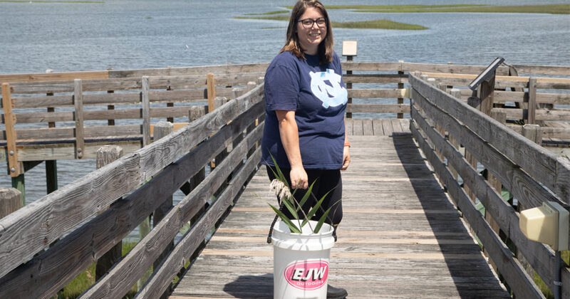 A woman stands on a pier holding a bucket. She is wearing a UNC tee-shirt.