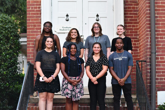 Eight students pose for a photo outside the Graduate Student Center; a brick building with white double doors.