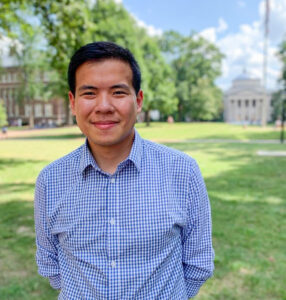 An Asian man in a blue checkered shirt stands smiling on the grass at Polk Place, with Wilson library just visible in the distance.