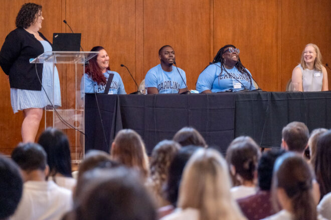 A woman stands at a podium holding a laptop. She is smiling at the table of panelists next to her. At the table, three students are wearing matching "Carolina" t-shirts. A fourth student is smiling and look back at the first three. 