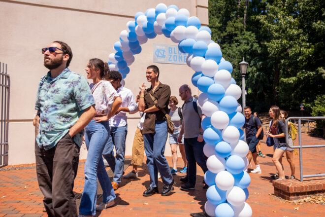 A number of students are walking through an arch made of blue and white balloons. They are dressed for hot weather and chatting with each other.